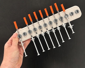 May include: A white plastic organizer tray holding nine clear plastic syringes with orange caps. The tray features circular cutouts for the syringes and is marked with the letters GL, L, S, and C, 2N. Each syringe has a white plunger and a clear barrel.