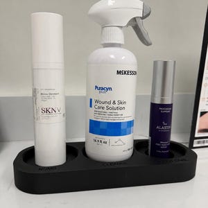 May include: A black rectangular organizer tray with three circular cutouts labeled Numb, Cleanse, and Post Care. It holds a white SKNV ointment bottle, a white McKesson Puracyn Plus wound and skin care solution spray bottle, and a purple Alastin serum.