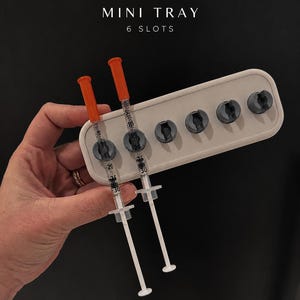 Könnte beinhalten: Ein weißes, rechteckiges Mini-Tablett mit sechs runden Aussparungen, von denen zwei durchsichtige Spritzen mit orangefarbenen Kappen und schwarzen Messmarkierungen enthalten. Der Text "THE SETUP COLLECTIVE" und "MINI TRAY 6 SLOTS" ist in Weiß über dem Tablett aufgedruckt.