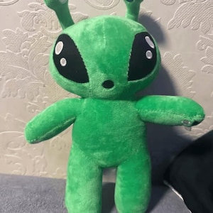 Può includere: Un peluche alieno verde brillante caratterizzato da grandi occhi ovali neri con riflessi bianchi, due antenne con punte circolari bianche e blu e un piccolo naso nero. La morbida creatura imbottita ha una testa arrotondata e arti semplici.