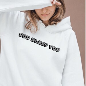 Peut inclure: Un sweat-shirt à capuche blanc avec le texte GOD BLESS YOU imprimé en lettres noires grasses et contourées sur la poitrine. Le vêtement possède des manches longues et une capuche, avec le texte positionné en diagonale sur le devant.
