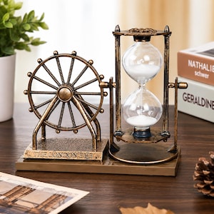 May include: A decorative bronze-colored metal ornament featuring a miniature Ferris wheel labeled THE LONDON EYE next to a rotating hourglass filled with white sand. The entire structure is mounted on a rectangular base with a metallic finish.