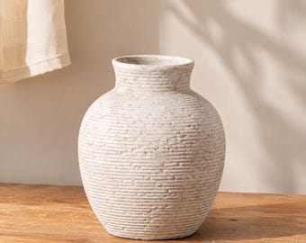 May include: A round, off-white vase with a textured, ribbed surface featuring horizontal lines and a weathered, stone-like finish. The vase has a wide, bulbous body that tapers toward a short, narrow neck with a simple, open circular rim.