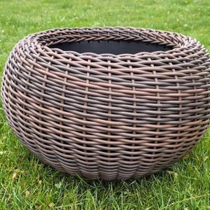 Puede incluir: Una maceta redonda de mimbre tejido en color marrón oscuro con un exterior texturizado similar a una cesta y un revestimiento interior negro liso. La maceta presenta un borde grueso trenzado y una forma bulbosa, diseñada para plantas.