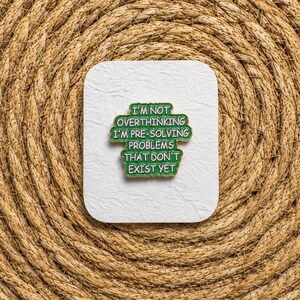 May include: A green enamel pin with gold-toned edges featuring the white text: I'M NOT OVERTHINKING I'M PRE-SOLVING PROBLEMS THAT DON'T EXIST YET. The pin is mounted on a square white card with rounded corners, centred on a textured woven surface.