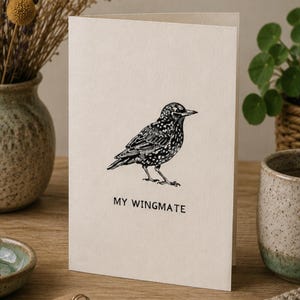 Może przedstawiać: Złożona kartka okolicznościowa z kremowego papieru. Z przodu znajduje się czarna ilustracja szpaka oraz napis "MY WINGMATE". Z tyłu widnieje wiersz o kluczu szpaków oraz tekst "© WrenMoonStudio".