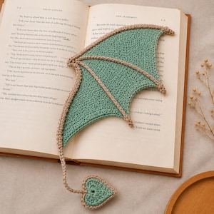 May include: A crochet bookmark shaped like a dragon wing with a heart-shaped tail. The wing is light green with tan trim, and the tail is a matching light green heart with tan edging. The bookmark is resting on an open book titled The Alchemist.