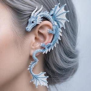 Può includere: Un ear cuff all'uncinetto a forma di drago, realizzato con filo azzurro e bianco e decorato con piccole perline bianche lungo i bordi. Il drago avvolge l'orecchio, con la testa rivolta verso l'alto e una lunga coda curva verso il basso.