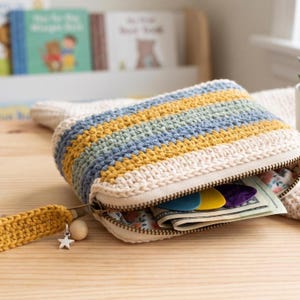 May include: A rectangular crochet zipped pouch featuring horizontal stripes in cream, mustard yellow, sage green, and dusty blue. The pouch has a matching mustard yellow wrist strap with a small silver star charm and a wooden bead attached.