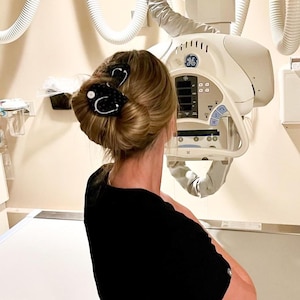Puede incluir: Una persona con cabello rubio recogido en un moño, asegurado con dos pinzas negras. Las pinzas presentan un diseño de estetoscopio blanco. La persona lleva una camisa negra y está en un entorno médico con una máquina de rayos X.