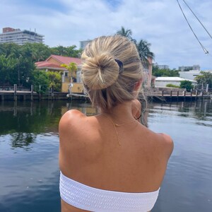 May include: A woman with blonde hair in a bun, wearing a white strapless top and a gold necklace, stands on a dock overlooking a canal. The water is calm and reflects the blue sky and white clouds.