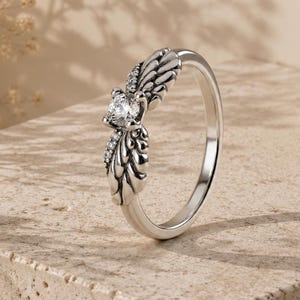 May include: A silver-toned ring featuring a central round clear gemstone set within a band designed to resemble two stylized wings. The metal surface displays intricate carved feather details and small embedded clear stones along the wing edges.