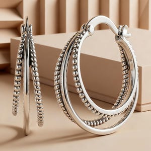May include: A pair of silver hoop earrings featuring a triple-layered design. Each earring consists of two textured beaded hoops and one smooth polished hoop, all connected by a hinged clasp mechanism at the top for secure closure.
