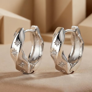 May include: A pair of silver hoop earrings featuring a wavy, textured surface. Each hoop is decorated with multiple engraved star motifs, with each star containing a small, clear, embedded stone at its center. The earrings have a polished finish.