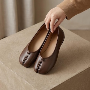 May include: A pair of dark brown leather-like ballet flats featuring a distinctive split-toe design. The shoes have a smooth, solid-coloured exterior with a small decorative knot detail at the centre of the vamp and a low-profile silhouette.