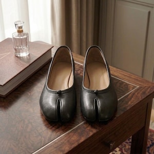 May include: A pair of dark gray leather tabi shoes featuring a distinct split-toe design, a low-cut topline, and a small decorative knot detail at the center of the vamp. The shoes have a smooth finish and a beige interior lining.