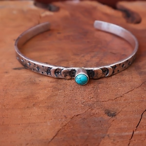 May include: A silver cuff bracelet featuring a central round turquoise stone. The band is engraved with a repeating pattern of moon phases and small four-pointed stars. The metal has a darkened, oxidized finish in the recessed areas of the design.