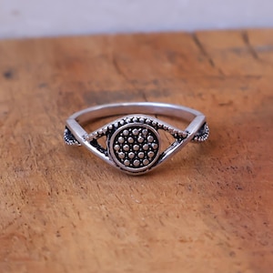 May include: A silver-toned ring featuring a central circular element with a textured, dotted surface. The band splits into two delicate, curved arms that frame the center, creating an eye-like shape. The metal has a dark, oxidized finish.