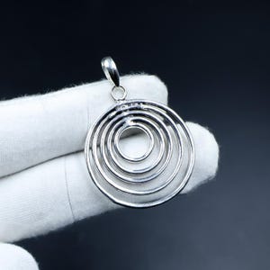 Op de afbeelding: Een zilverkleurige hanger bestaande uit vier concentrische cirkels van verschillende groottes, die aan de bovenkant zijn verbonden met een kleine, gepolijste hangerlus. Het metaal heeft een gladde, reflecterende afwerking en het ontwerp bestaat volledig uit open cirkelvormige frames.