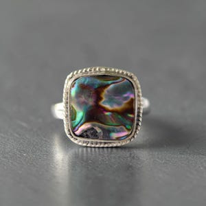 May include: A square-shaped ring featuring an iridescent abalone shell inlay with swirling patterns of green, purple, and blue. The stone is set within a silver-toned metal frame that has a decorative twisted rope texture along the outer edge.