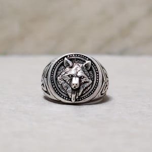 May include: A silver-toned metal ring featuring a detailed relief of a wolf head centered within a circular frame. The band of the ring is decorated with intricate geometric knotwork patterns, and the metal has a polished finish with darkened recessed areas to highlight the sculpted features of the wolf.