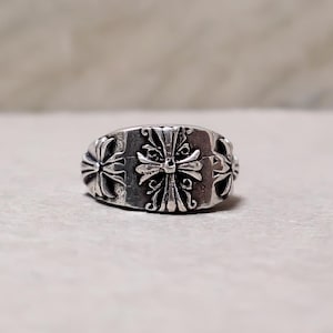 May include: A silver ring featuring a band with three embossed cross motifs. The central cross is larger and more detailed, flanked by two smaller crosses on either side. The metal has a darkened, oxidised finish in the recessed areas to highlight the intricate scrollwork and raised cross designs.
