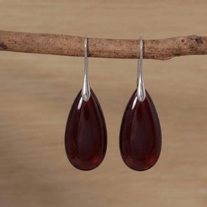 May include: A pair of drop earrings featuring smooth, deep red, teardrop-shaped stones suspended from polished silver-toned metal hooks. The earrings hang vertically, showcasing a translucent, dark crimson hue and a simple, clean design.