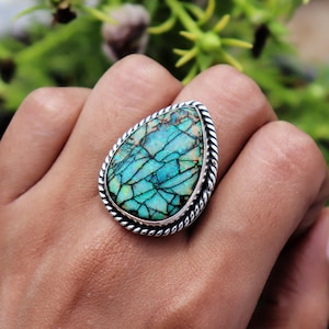May include: A teardrop-shaped ring featuring a vibrant turquoise and black matrix stone set in a polished silver frame. The setting is accented with a detailed twisted rope texture along the outer edge, creating a distinct border around the gemstone.