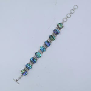 May include: A link bracelet featuring ten oval-shaped abalone shell inlays with iridescent blue, green, and purple tones. The shell pieces are set in silver-toned metal frames connected by small bars, ending with a toggle clasp and extender chain.