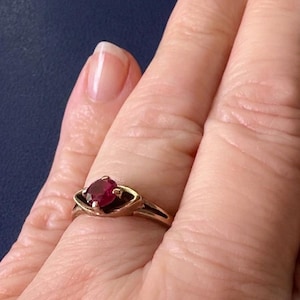 May include: A gold ring featuring a single oval-cut deep red gemstone set in a prong setting. The band has a smooth, polished finish and a unique design that tapers towards the centre, framing the central stone with curved metal accents.