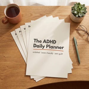 Może przedstawiać: Stos białych kartek papieru z tytułem The ADHD Daily Planner i podtytułem undated brain-friendly zero guilt. Planerowi towarzyszy żebrowany brzoskwiniowy kubek, mały zielony sukulent w doniczce, zielony długopis oraz okulary w cienkich oprawkach.