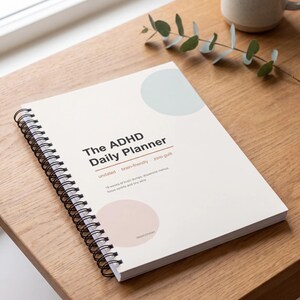 May include: A spiral-bound daily planner with a white cover featuring the text The ADHD Daily Planner, undated, brain-friendly, zero guilt, and 18 weeks of brain dumps, dopamine menus, focus sprints and tiny wins, with light blue and pink circles.