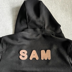 May include: A black hooded sweatshirt featuring a textured, ribbed fabric finish. The back of the garment displays the name SAM in large, rounded, light brown, leather-like applique letters centred horizontally across the upper back area.