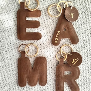 Op de afbeelding: Vier bruine leren sleutelhangers in de vorm van de letters E, A, M en R. De A-sleutelhanger heeft de naam AZITA en een Ram-sterrenbeeld op een rond label. De R-sleutelhanger bevat een rond label met een Weegschaal-symbool. Gouden ringen.