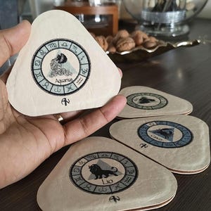 May include: A set of triangular off-white coasters featuring embroidered zodiac symbols. Each coaster displays a circular zodiac wheel with a central illustration of a sign, including Aquarius, Cancer, and Leo, with text labels below.