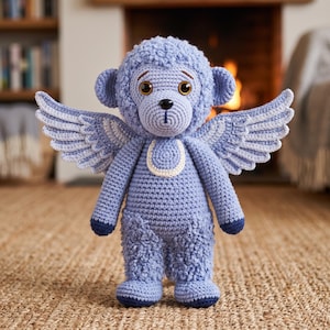 May include: A light blue crocheted monkey plush toy featuring large brown eyes, a small black nose, and a textured curly head and legs. The monkey has two large, detailed crocheted wings extending from its back and a cream-colored chest patch.