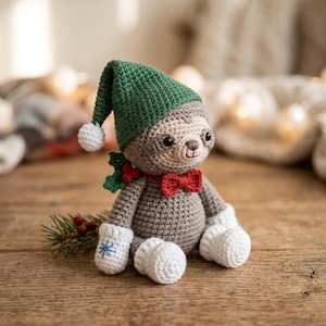 Può includere: Un bradipo di peluche all'uncinetto che indossa un cappello a punta verde con un pompon bianco, un papillon rosso e muffole bianche con un ricamo a fiocco di neve blu. Il bradipo ha pelliccia marrone, occhi neri e un rametto di agrifoglio.