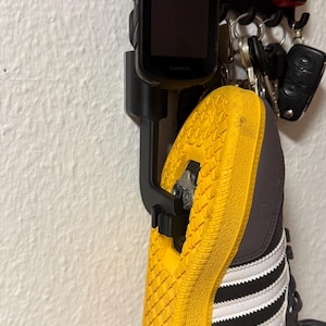 Könnte beinhalten: Ein schwarzer Wandhalter mit einem Garmin-Fahrradcomputer und einem grauen Radschuh mit leuchtend gelber Sohle. Der Halter verfügt über einen Haken für Schlüssel und eine kleine rote Olight-Taschenlampe. Der Schuh ist mit einem Metall-Cleat befestigt.