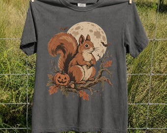 May include: A charcoal gray short-sleeve t-shirt featuring a graphic print of a brown squirrel perched on a tree branch. The design includes a large full moon, a small orange jack-o'-lantern, autumn leaves, and two flying bats in the background.