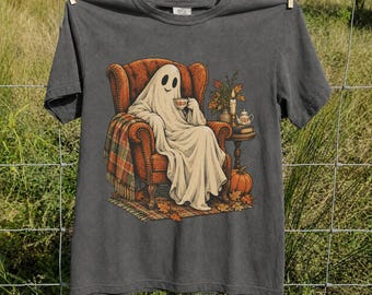 May include: A charcoal gray t-shirt featuring a graphic of a ghost draped in a white sheet, sitting in a brown armchair while holding a teacup. The illustration includes a plaid blanket, a small side table with a teapot, and a pumpkin.