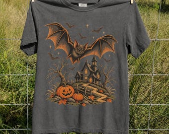 May include: A charcoal gray t-shirt featuring a graphic print of a large bat flying over a haunted house, bare trees, and two orange pumpkins. The illustration includes small stars and autumn leaves, rendered in shades of orange and brown.