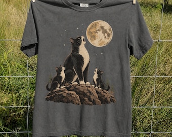 May include: A charcoal gray short-sleeve t-shirt featuring a graphic print of a large black and white cat and two smaller kittens sitting on a rocky outcrop, all looking up at a large, textured full moon surrounded by small white stars.