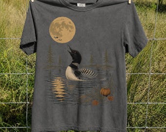 May include: A charcoal gray short-sleeve t-shirt featuring a graphic print of a loon swimming on a lake under a large full moon. The design includes silhouettes of pine trees, a small pumpkin on the shore, and a reflection of the moon on the water.