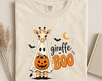 May include: A cream-colored t-shirt featuring a graphic of a giraffe wearing a white ghost costume and holding a jack-o'-lantern bucket filled with candy. The design includes two black bats, a yellow crescent moon, and the text giraffe BOO.