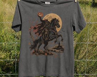 May include: A charcoal gray short-sleeve t-shirt featuring a graphic print of a headless horseman riding a black horse against a large yellow moon. The rider holds a carved orange pumpkin, and the scene includes a dark silhouette of a building.