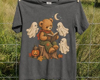 May include: A charcoal gray t-shirt featuring a graphic of a brown teddy bear sitting on a tree stump, reading an open book. The bear is surrounded by three white ghosts, a jack-o'-lantern, a crescent moon, and scattered autumn leaves.