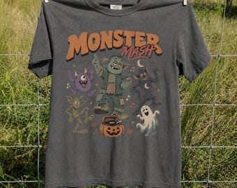 May include: A charcoal gray short-sleeve t-shirt featuring the text "MONSTER MASH" in orange. The graphic includes a central green monster, a purple one-eyed creature, a ghost, and a dark figure dancing around a jack-o'-lantern record player.