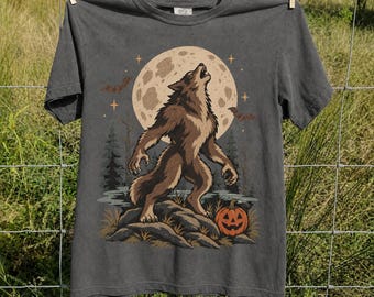 May include: A charcoal gray short-sleeve t-shirt featuring a graphic print of a brown werewolf howling at a large beige full moon. The design includes silhouettes of pine trees, flying bats, small stars, and an orange jack-o'-lantern pumpkin.
