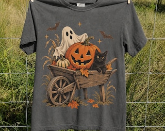 May include: A charcoal gray short-sleeve t-shirt featuring a graphic print of a wooden wheelbarrow filled with a large orange jack-o'-lantern, a small striped pumpkin, a white ghost, and a black cat, surrounded by autumn leaves and bats.
