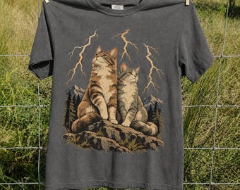 May include: A charcoal gray short-sleeve t-shirt featuring a graphic print of two tabby cats sitting on a rocky outcrop. The background displays a mountain landscape with pine trees and yellow lightning bolts striking from the sky.